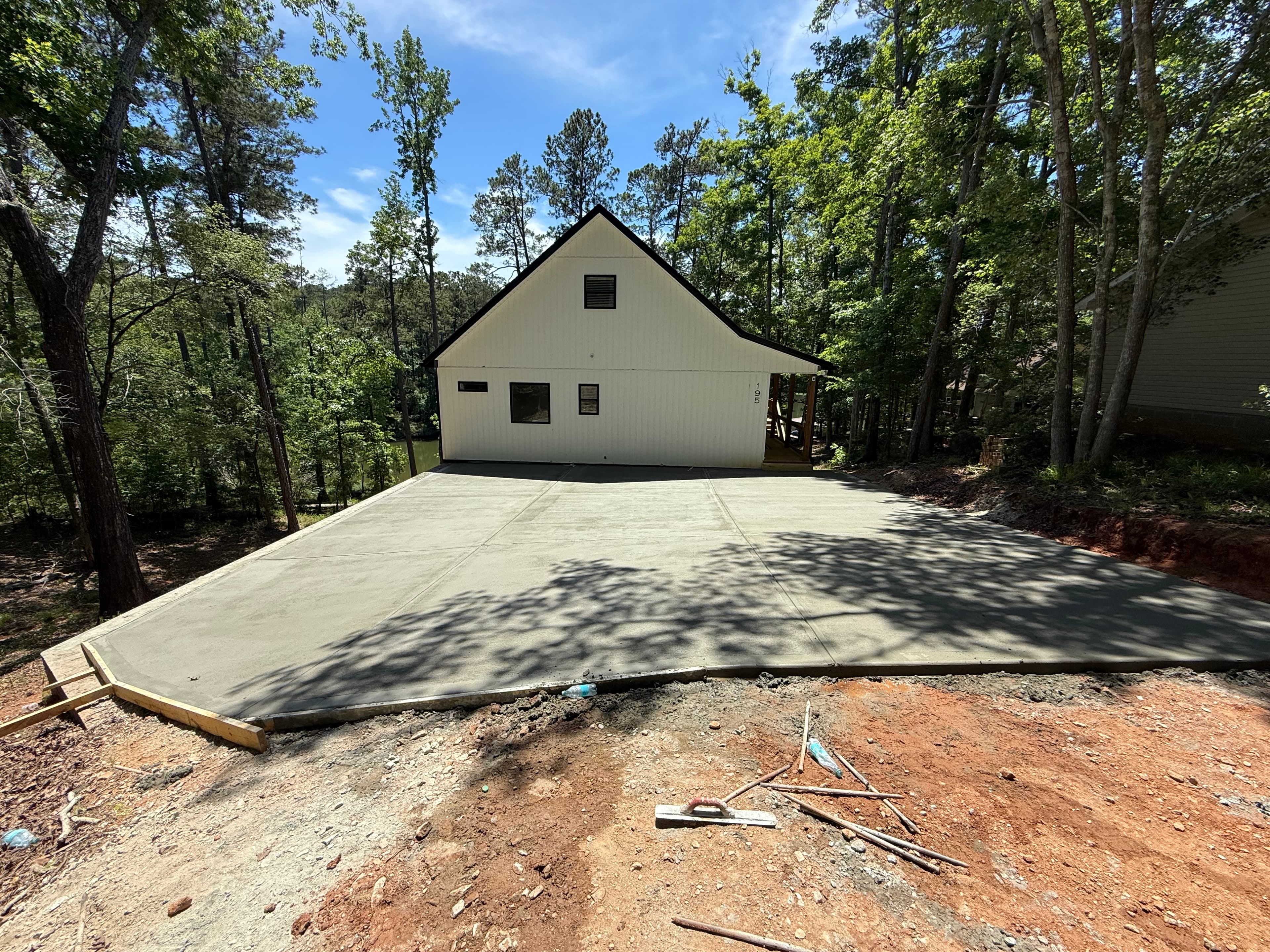 White modern house with a large newly poured concrete driveway surrounded by lush green trees.