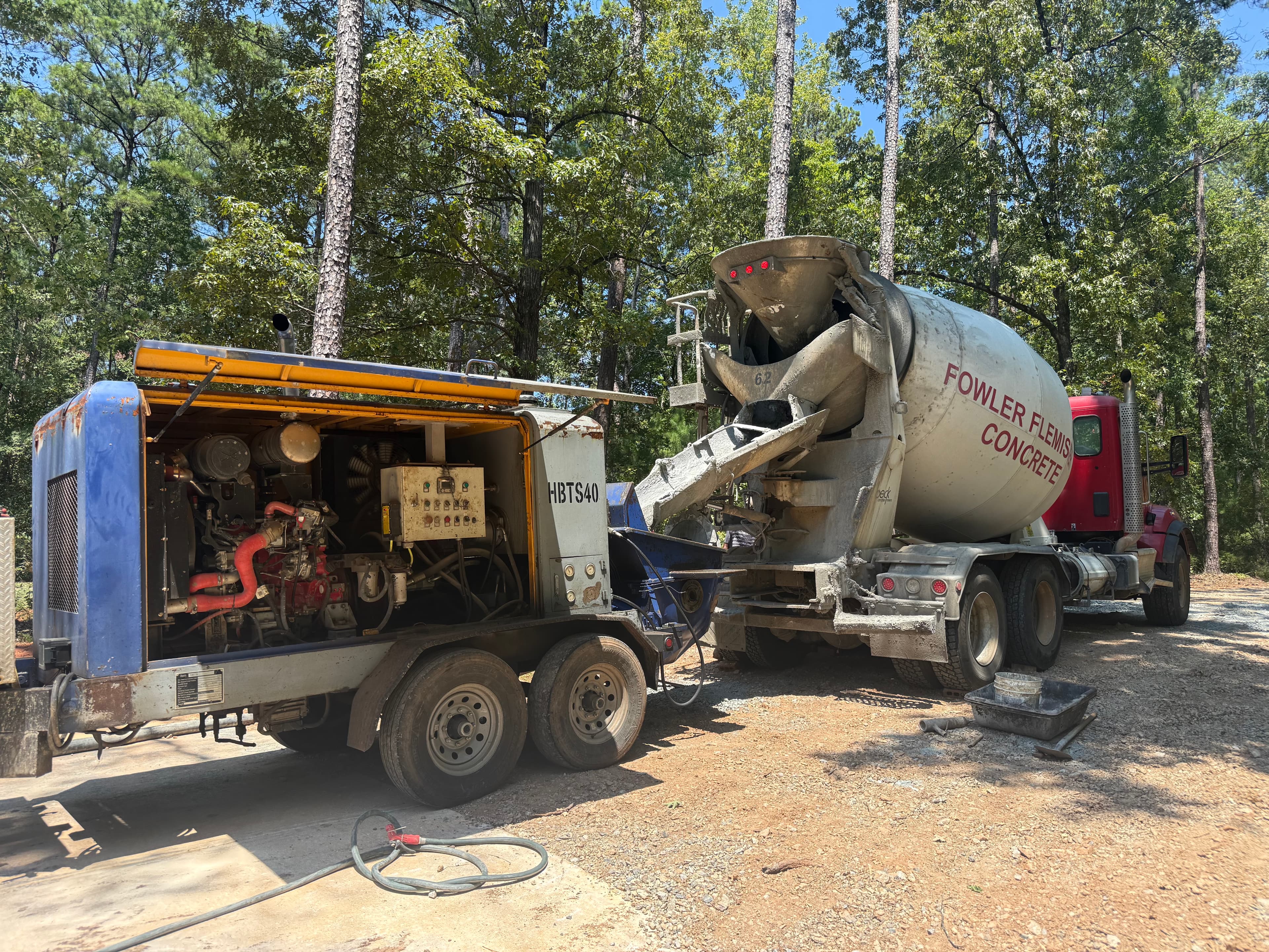 Concrete mixer truck pouring wet concrete into a trailer-mounted pump at a wooded construction site.