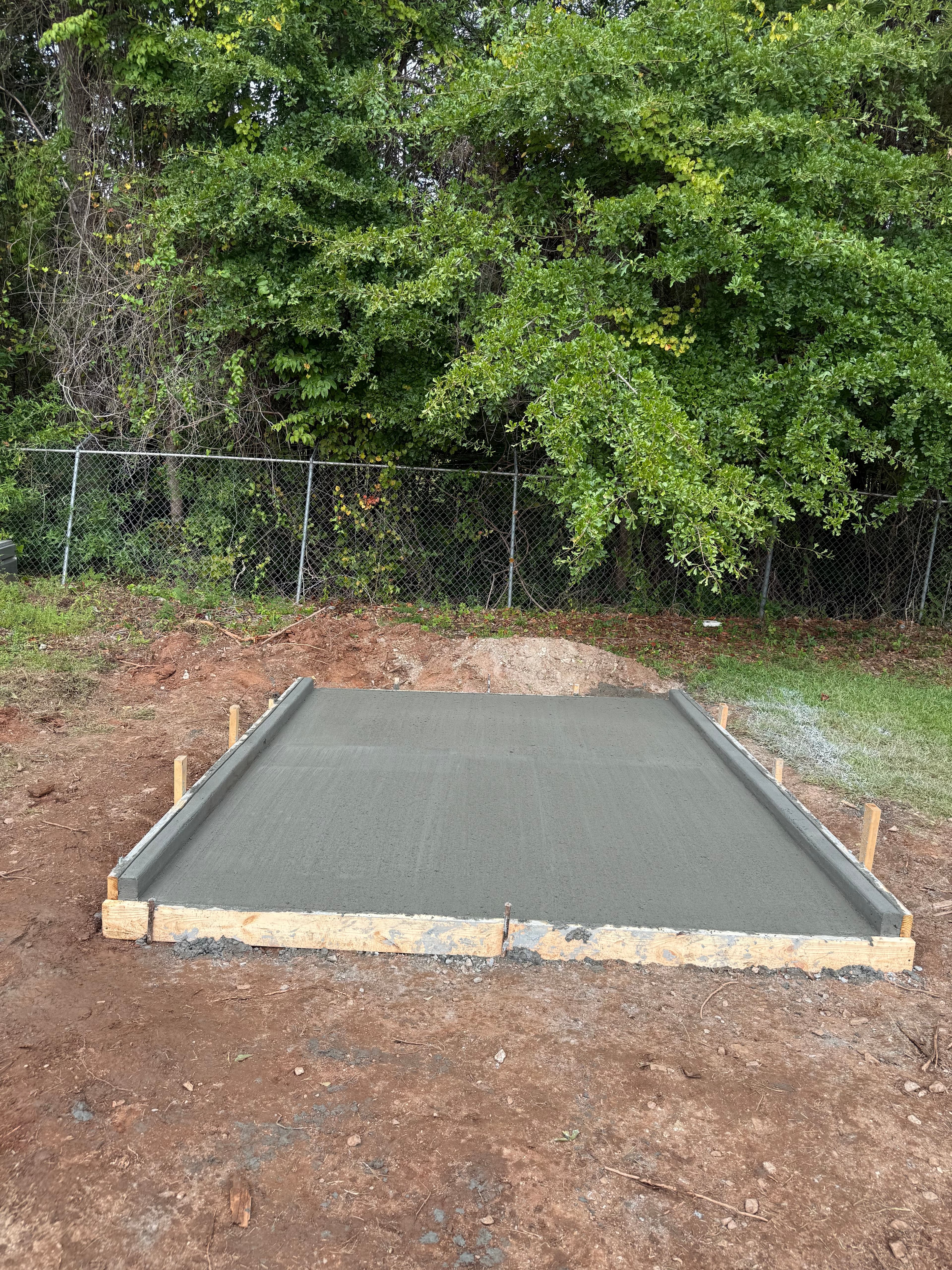 Freshly poured concrete slab with raised side curbs in wooden forms on dirt ground.