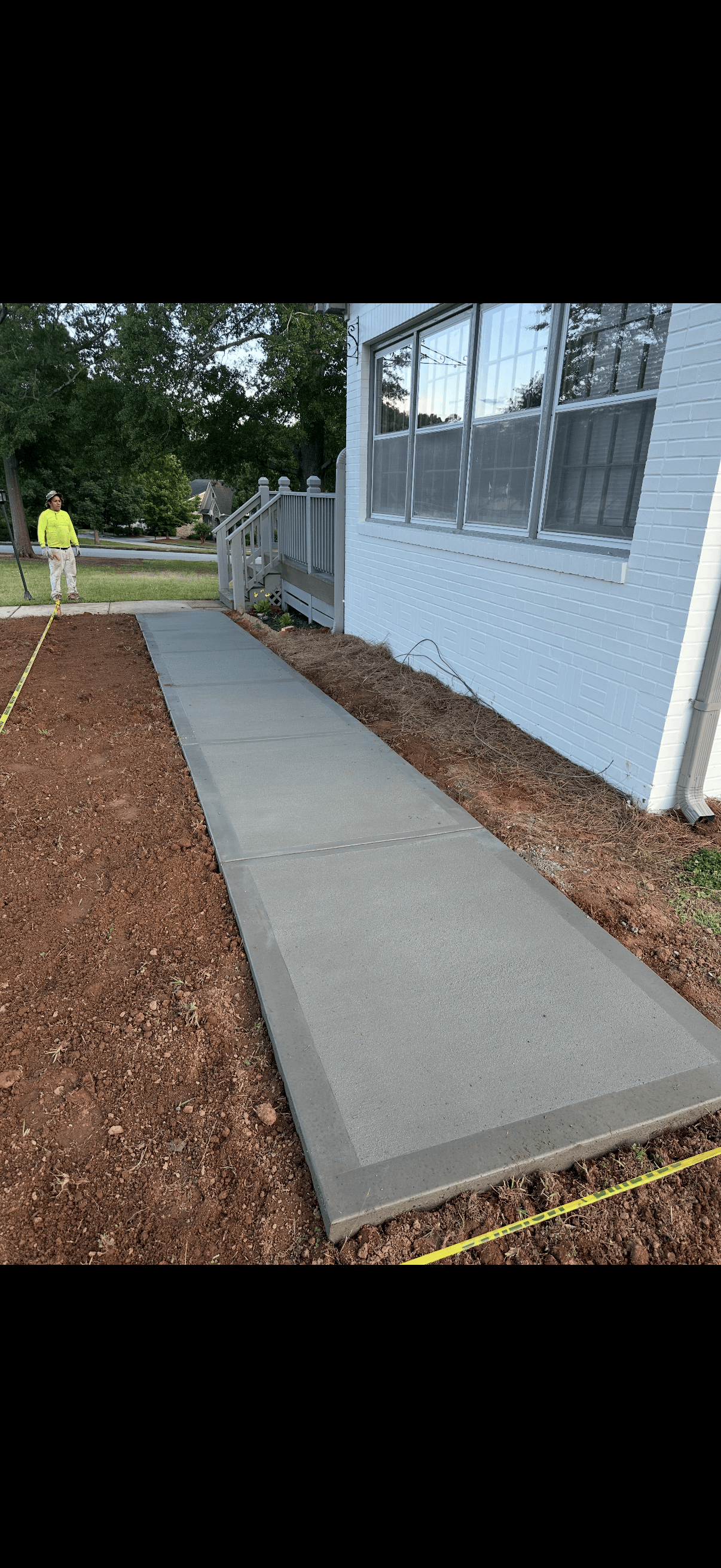 Newly finished concrete walkway alongside a white brick house with red dirt and caution tape.