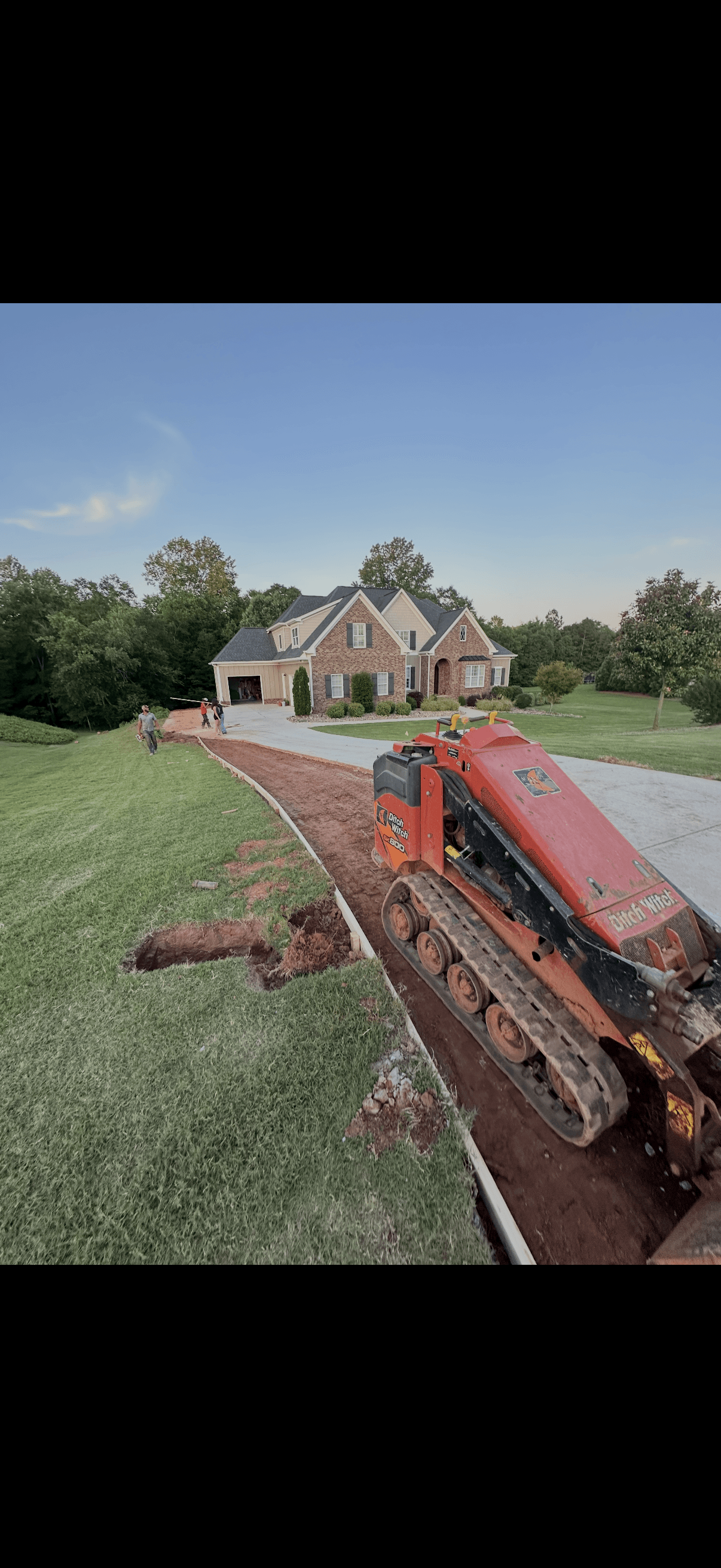 Red Ditch Witch mini skid steer preparing a dirt path by a large brick house.